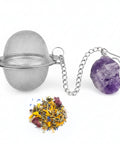 Ein silberner, runder Teeball-Filter aus Drahtgeflecht ist über eine Kette mit einem rohen, violetten Amethystkristall verbunden. Unterhalb des Teeballs liegt ein kleiner Haufen bunter loser Kräutertee, der gelbe, blaue und rote getrocknete Blütenblätter enthält, alles vor einem weißen Hintergrund.