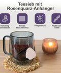 Teesieb mit Rosenquarz-Anhänger in einer Tasse neben einer Kerze.