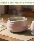 Eine dampfende rosa Keramikschale mit schaumigem grünen Matcha-Tee auf einem Tisch neben einem Bambuslöffel und Besenhalter, darüber der Text „Genieße den Matcha-Moment“.