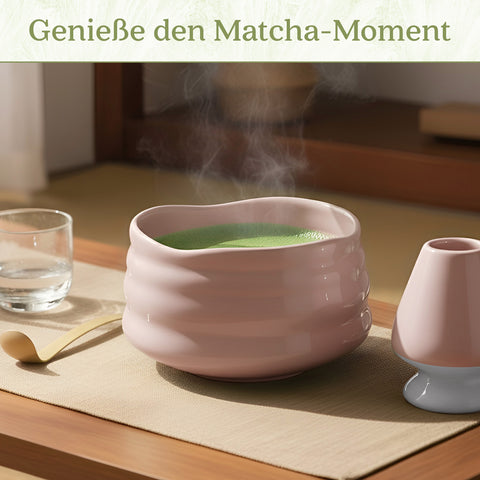 Eine dampfende rosa Keramikschale mit schaumigem grünen Matcha-Tee auf einem Tisch neben einem Bambuslöffel und Besenhalter, darüber der Text „Genieße den Matcha-Moment“.