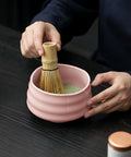 Eine Nahaufnahme einer Person, die mit einem traditionellen Bambusbesen (Chasen) Matcha-Tee in einer rosa Keramikschale auf einem dunklen Holztisch anrührt.
