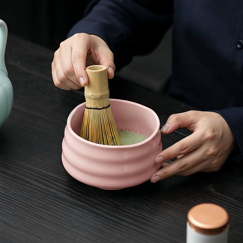 Eine Nahaufnahme einer Person, die mit einem traditionellen Bambusbesen (Chasen) Matcha-Tee in einer rosa Keramikschale auf einem dunklen Holztisch anrührt.