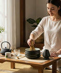 Gewurzladen Nafels Frau bereitet Matcha Tee mit Besen und schwarzer Schale auf einem Holztisch zu.