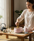 Eine Frau sitzt ruhig an einem niedrigen Holztisch und bereitet mit einem Bambusbesen Matcha in einer rosa Schale zu, umgeben von einem traditionellen Tee-Set.