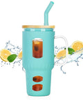 Großer Glasbecher mit mintgrüner Silikonhülle, Holzdeckel, Griff und gebogenem Trinkhalm, gefüllt mit Eistee, umgeben von spritzenden Zitronenscheiben und Minze.