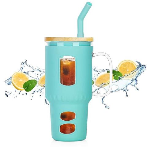 Großer Glasbecher mit mintgrüner Silikonhülle, Holzdeckel, Griff und gebogenem Trinkhalm, gefüllt mit Eistee, umgeben von spritzenden Zitronenscheiben und Minze.
