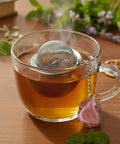 Dampfende Tasse Tee mit Netz-Teesieb und rotem Kristall-Anhänger auf einer Holzoberfläche.