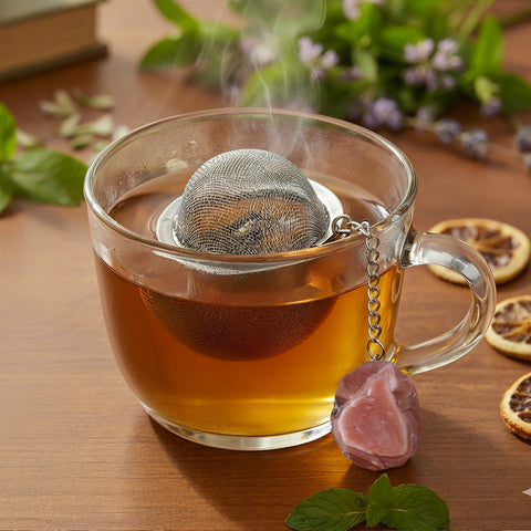 Dampfende Tasse Tee mit Netz-Teesieb und rotem Kristall-Anhänger auf einer Holzoberfläche.