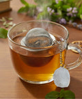 Dampfende Tasse Tee mit Netz-Teesieb und Kristall-Anhänger auf einer Holzoberfläche.