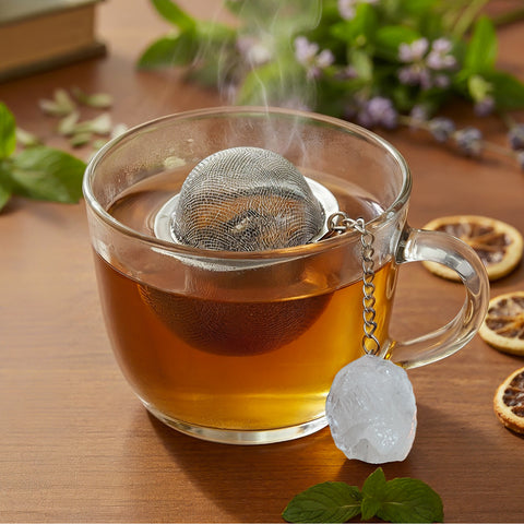 Dampfende Tasse Tee mit Netz-Teesieb und Kristall-Anhänger auf einer Holzoberfläche.
