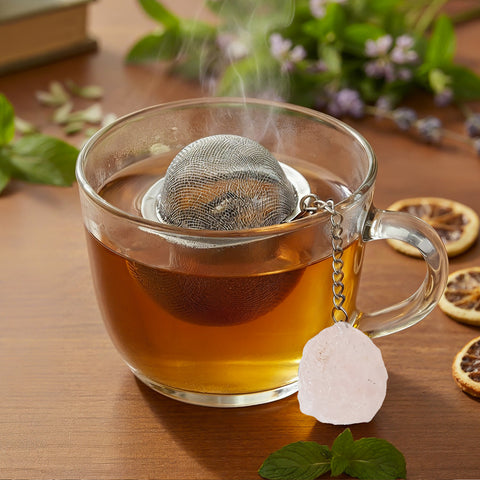 Nahaufnahme einer dampfenden Teetasse mit Sieb und Rosenquarz-Anhänger.