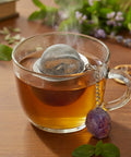 Nahaufnahme einer dampfenden Teetasse mit Sieb und Amethyst-Kristallanhänger.