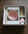 Eine weiße Geschenkbox auf einer Holzoberfläche, die eine rosa Keramikschale, einen Bambuslöffel, einen Bambusbesen und einen rosa Besenhalter enthält.