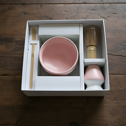 Eine weiße Geschenkbox auf einer Holzoberfläche, die eine rosa Keramikschale, einen Bambuslöffel, einen Bambusbesen und einen rosa Besenhalter enthält.