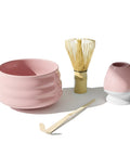 Ein traditionelles rosa Matcha-Tee-Set bestehend aus einer geriffelten Keramikschale, einem Bambusbesen (Chasen), einem Bambuslöffel (Chashaku) und einem rosa Besenhalter auf weißem Hintergrund.