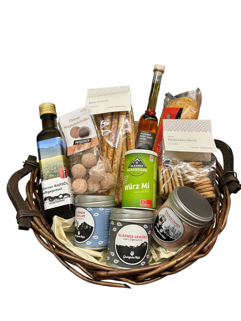 Ein dunkler Weidenkorb gefüllt mit Schweizer Spezialitäten aus Glarus, darunter eine Flasche Glarner Rapsöl, eine Flasche Kräuter-Spirituose, Packungen mit salzigem Gebäck, Schokoladentrüffeln und Dosen mit Glarner Gewürzen.