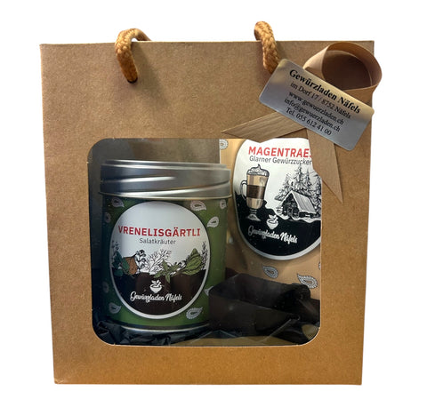 Eine braune Geschenktüte vom „Gewürzladen Näfels“ mit einer Dose „Vrenelisgärtli“ Salatkräuter und einer Packung „Magentraes“ Gewürzzucker, sichtbar durch ein Sichtfenster.