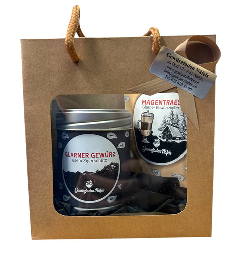 Eine braune Geschenktüte vom „Gewürzladen Näfels“ mit einer Dose „Glarner Gewürz“ und einer Packung „Magentraes“ Gewürzzucker, versehen mit einem Sichtfenster und einem silbernen Kärtchen.