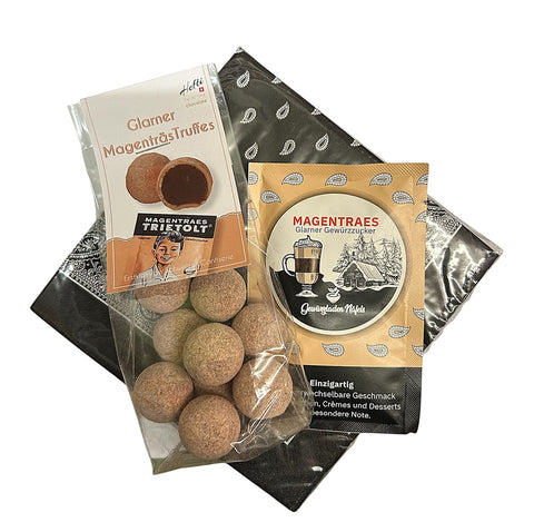 Ein Klarsichtbeutel mit „Glarner Magenträs-Truffes“ von Hefti und ein Beutel Magenträs-Gewürzzucker vom Gewürzladen Näfels, angerichtet auf schwarzem Geschenkpapier mit Paisley-Muster.