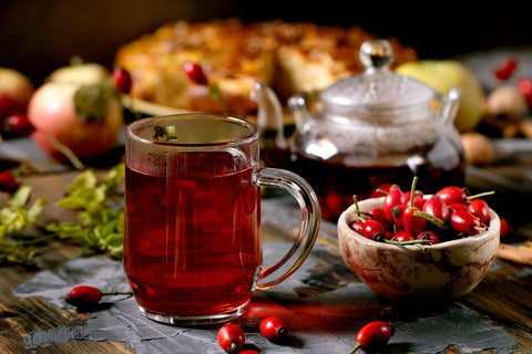 Eine Glastasse mit rotem Früchtetee auf einem rustikalen Holztisch, umgeben von frischen Hagebutten, einer Glasteekanne und einem verschwommenen Kuchen im Hintergrund.