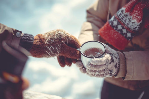 Nahaufnahme von zwei Personen in Winterkleidung und Strickhandschuhen, die sich im Freien im Schnee eine dampfende Emailletasse Tee teilen.