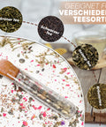 Ein Glas-Teesieb in Reagenzglasform liegt auf einem Teller mit verschütteten Teeresten. Es werden Kreise mit losem Grünem Tee, Schwarztee, Oolong-Tee und Kräutertee als Beispiele für verschiedene Teesorten gezeigt.