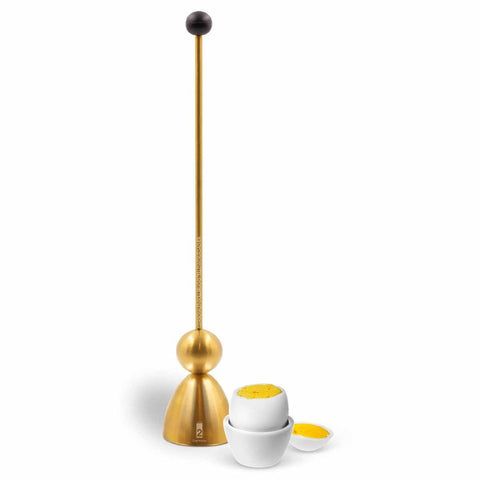 Ein hoher, goldener Messing-Eierschalensollbruchstellenverursacher „Clack“ neben einem Ei im Eierbecher.
