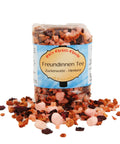 Eine Klarsichtpackung „Freundinnen Tee“, die hinter einem ausgestreuten Haufen der farbenfrohen Früchteteemischung mit rosa Marshmallows, getrockneten Beeren und Fruchtstücken steht.