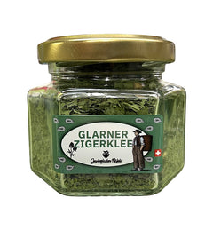 Ein kleines, quadratisches Glas mit goldfarbenem Deckel, gefüllt mit getrocknetem, grünem "Glarner Zigerklee" (Schabzigerklee). Das Etikett zeigt einen Schweizer Senn und die Aufschrift "Gewürzladen Näfels".