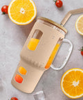 Ein mit Orangensaft gefüllter, beige-farbener Glasbecher mit Henkel und Strohhalm, umgeben von aufgeschnittenen Orangen, Zitronen, Kirschtomaten, Eiswürfeln und Wassertropfen auf einer grauen Oberfläche.