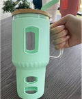 Eine Hand hält einen großen Glasbecher mit hellgrüner (mintgrüner) Silikonhülle, Holzdeckel und Glasgriff auf einem Tisch, mit weiteren Bechern im Hintergrund.
