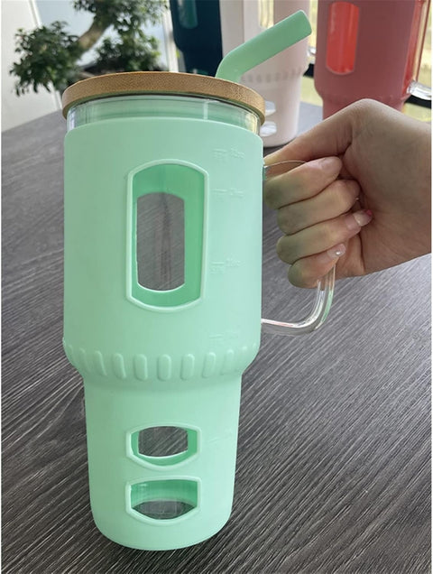 Eine Hand hält einen großen Glasbecher mit hellgrüner (mintgrüner) Silikonhülle, Holzdeckel und Glasgriff auf einem Tisch, mit weiteren Bechern im Hintergrund.