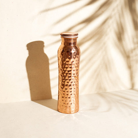 Eine gehämmert aussehende Kupferwasserflasche im Sonnenlicht mit Schatten von tropischen Blättern.