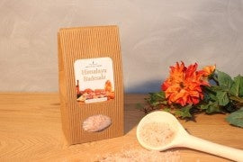 Ein brauner, gerippter Papierbeutel „Himalaya Badesalz“ neben einer weißen Schaufel mit rosa Salzkristallen und orangefarbenen Dekorationsblumen.