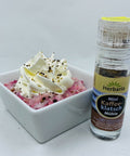 Eine kleine Herbaria „Mini Kaffeeklatsch Mühle“ neben einer weißen Schale mit rosa Dessert, garniert mit einer Schlagsahnehaube und einer Prise der Gewürzmischung.