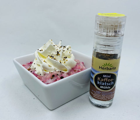 Eine kleine Herbaria „Mini Kaffeeklatsch Mühle“ neben einer weißen Schale mit rosa Dessert, garniert mit einer Schlagsahnehaube und einer Prise der Gewürzmischung.