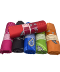 Ein Stapel farbenfroher, aufgerollter Strandtücher in Pink, Schwarz, Blau, Grün und Orange, mit einem roten Tuch obenauf; alle zeigen ein klassisches Paisley-Muster und Papierbanderolen.