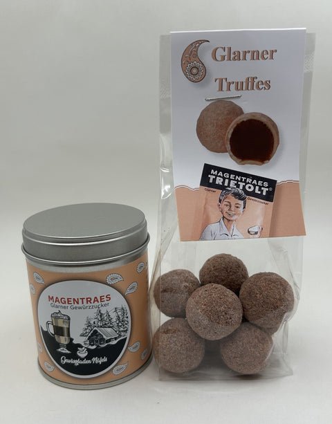 Eine silberne Blechdose "Magentraes Glarner Gewürzzucker" steht neben einem durchsichtigen Beutel mit mehreren Schokoladen-Trüffeln. Der Beutel ist mit "Glarner Truffes" und "Magentraes Trietolt Gewürzzucker" beschriftet.