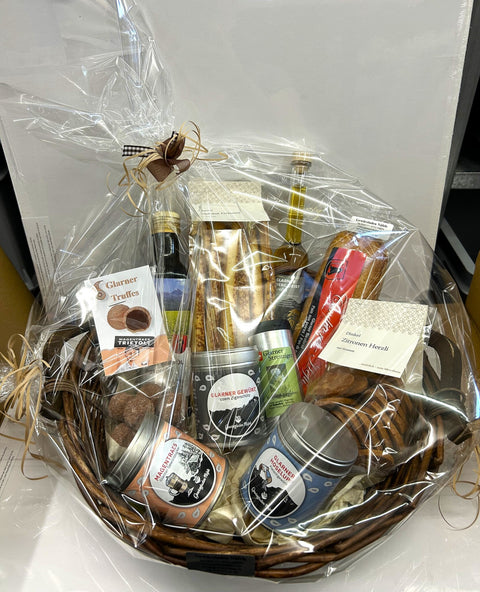 Ein Weiden-Geschenkkorb gefüllt mit Schweizer Spezialitäten aus Glarus, darunter Öle, Spirituosen, verschiedene Kekse und Trüffel sowie Gewürzdosen, alles fest in Klarsichtfolie mit Schleifen verpackt.