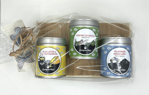 Drei etikettierte Gewürzdosen auf einem kleinen Holzbrett, in Klarsichtfolie verpackt mit Schleife und Schmetterlingsdeko.
