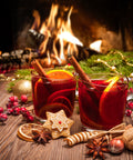Zwei Gläser roter Glühwein mit Zimtstangen und Orangenscheiben auf einem Holztisch, umrahmt von festlicher Dekoration und einem gemütlichen Kaminfeuer im Hintergrund.