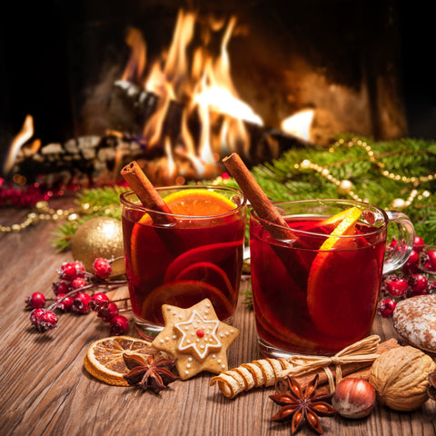 Zwei Gläser roter Glühwein mit Zimtstangen und Orangenscheiben auf einem Holztisch, umrahmt von festlicher Dekoration und einem gemütlichen Kaminfeuer im Hintergrund.