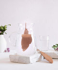 Kupferfeder-Infuser in einem Glaskrug neben einer zweiten Feder, einem Glas Wasser und Blumen.