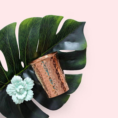 Ein handgehämmerter Kupferbecher mit Hammerschlag-Struktur liegt auf einem großen grünen Monstera-Blatt neben einer hellblauen Keramikblüte vor einem zartrosa Hintergrund.