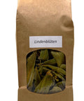 Ein brauner Papierbeutel mit einem kleinen Sichtfenster, das getrocknete, gelbgrüne Lindenblüten und Hochblätter zeigt, beschriftet mit einem weißen Etikett „Lindenblüten“.