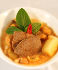 Nahaufnahme eines Massaman Currys mit Rindfleisch, Kartoffeln, Cashewkernen, garniert mit Thai-Basilikum und einer roten Chili-Scheibe in einer weißen Schüssel.