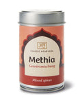 Eine silberne zylindrische Dose „Methia Gewürzmischung“ von Classic Ayurveda mit einem orange-roten Etikett, einem bogenförmigen Design und einer Aufschrift im Sanskrit-Stil.