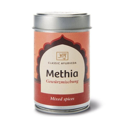 Eine silberne zylindrische Dose „Methia Gewürzmischung“ von Classic Ayurveda mit einem orange-roten Etikett, einem bogenförmigen Design und einer Aufschrift im Sanskrit-Stil.