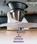 Der graue mixcover Thermo-Griff wird als Ständer genutzt, um den durchscheinenden Mixtopfdeckel stabil auf einer Holzarbeitsplatte abzustellen. Eine Küchenmaschine steht im verschwommenen Hintergrund. Text: „Sicheres Abstellen“.