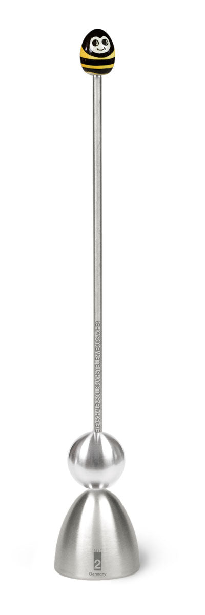 Ein Edelstahl-Eierschalensollbruchstellenverursacher „Clack“ von Take2 Germany mit Führungsstange und glockenförmigem Unterteil, abgeschlossen durch einen Keramikkopf im Design einer lächelnden schwarz-gelben Biene.